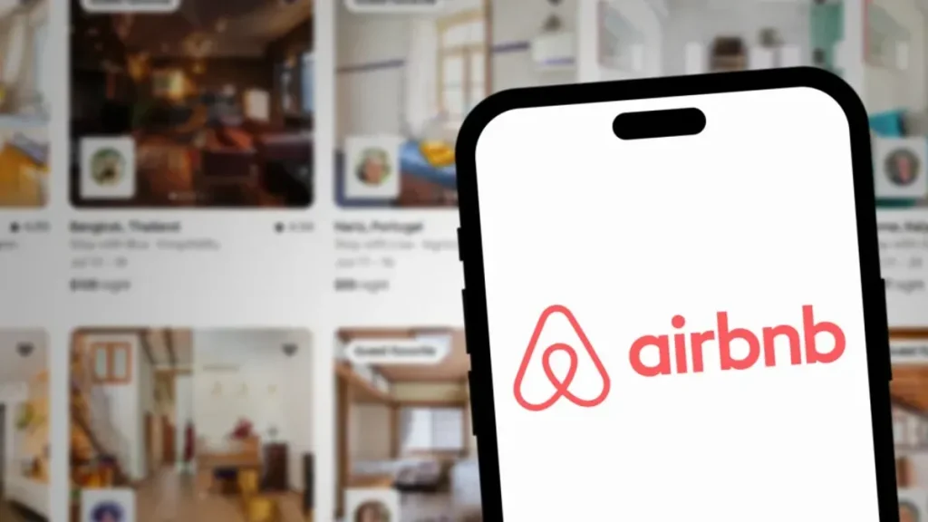 Aplicativo do Airbnb exibindo opções de hotéis, aluguel de carros e delivery de supermercado em um smartphone, com fundo de viagem.