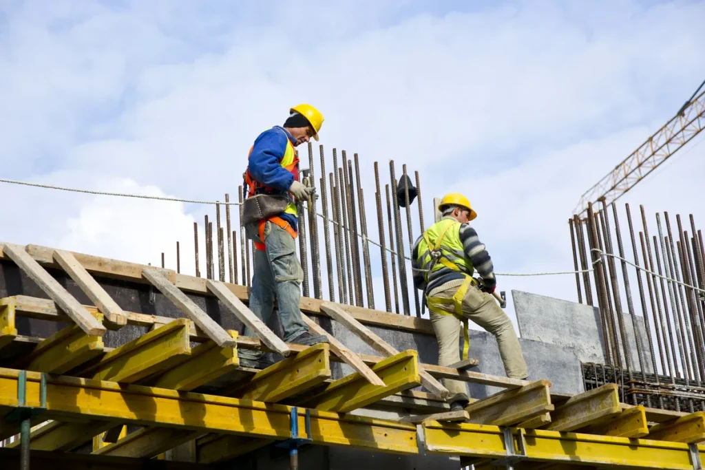 Trabalhadores da construção civil em canteiro de obras, discutindo planos, com elementos que sugerem aumento de custos no setor imobiliário.